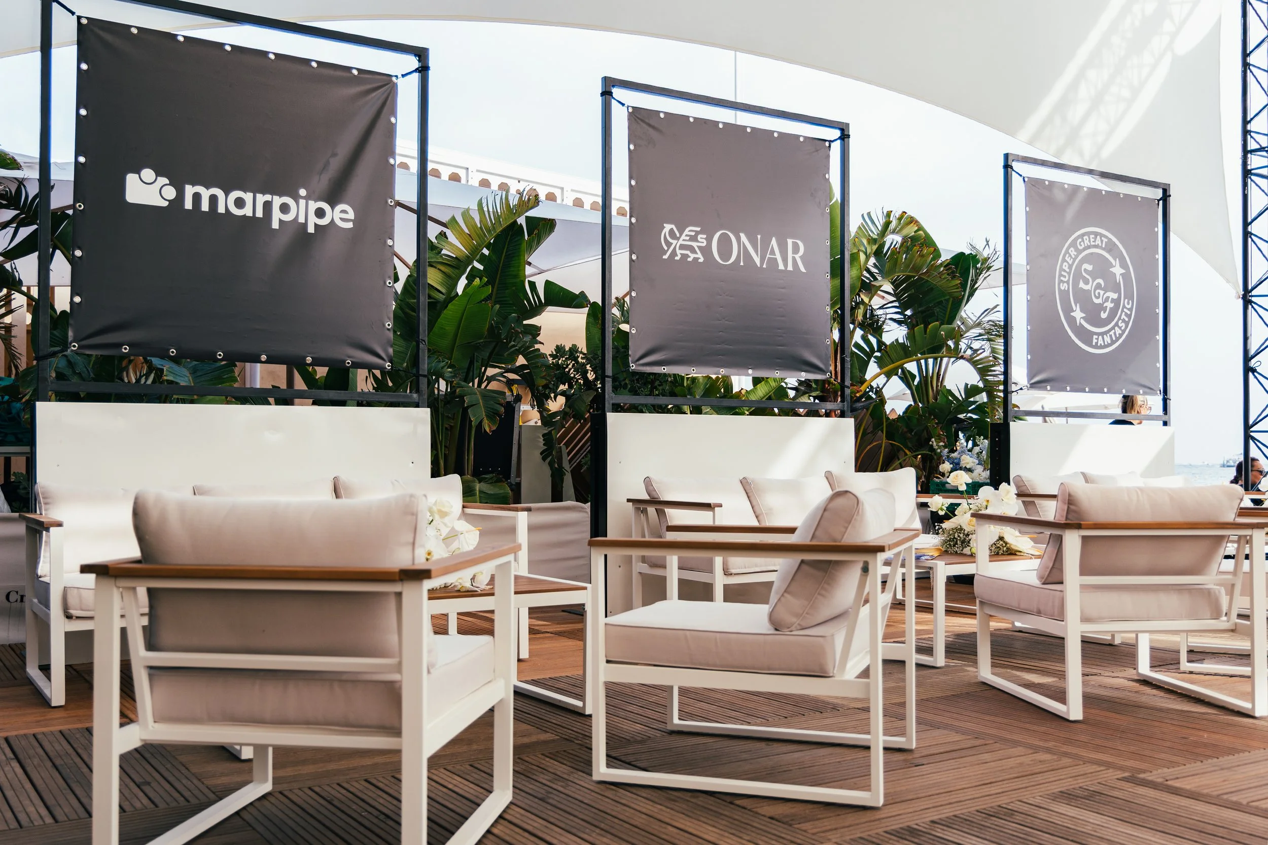 Outdoor seating area with white cushioned chairs and wooden tables, surrounded by green tropical plants, banners with logos of Marpipe, Onar, and Super Great Fantastic, under a white canopy.