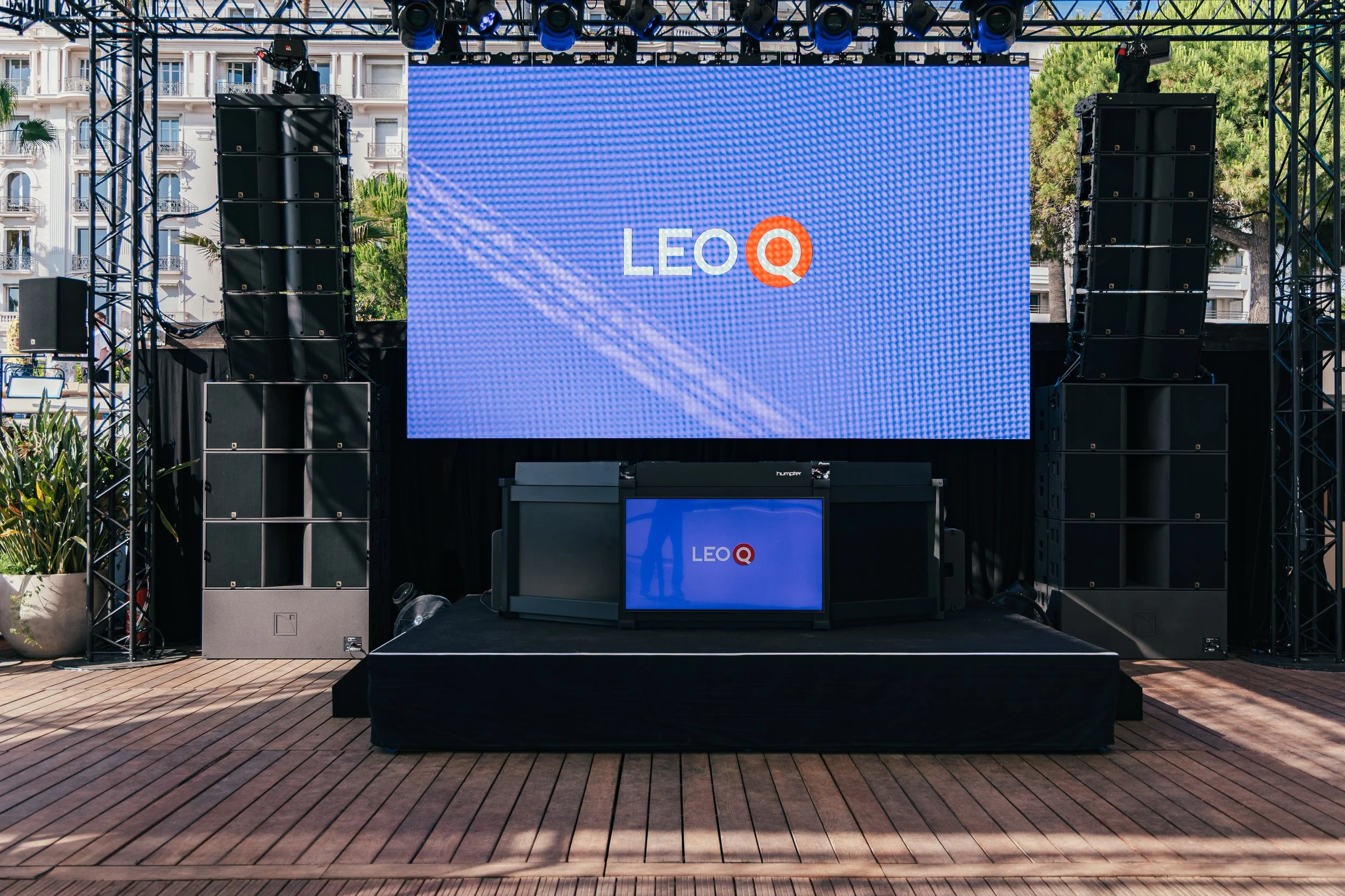 Outdoor stage with large LED screen displaying the logo 'LEO Q', surrounded by black speakers and equipment, set up on a wooden platform with a cityscape background.