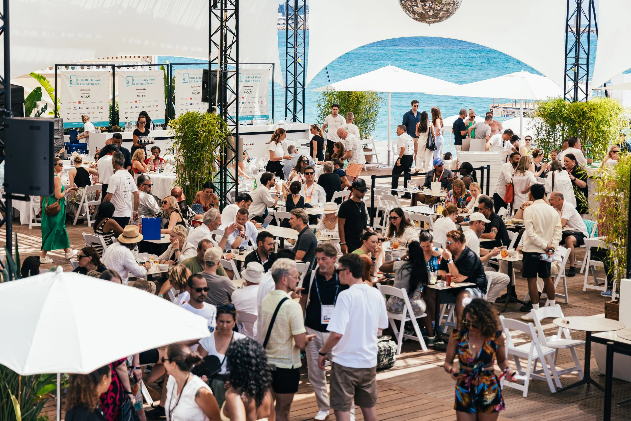 Crowd of people at outdoor event near the water with tables, chairs, umbrellas, and a stage in the background.