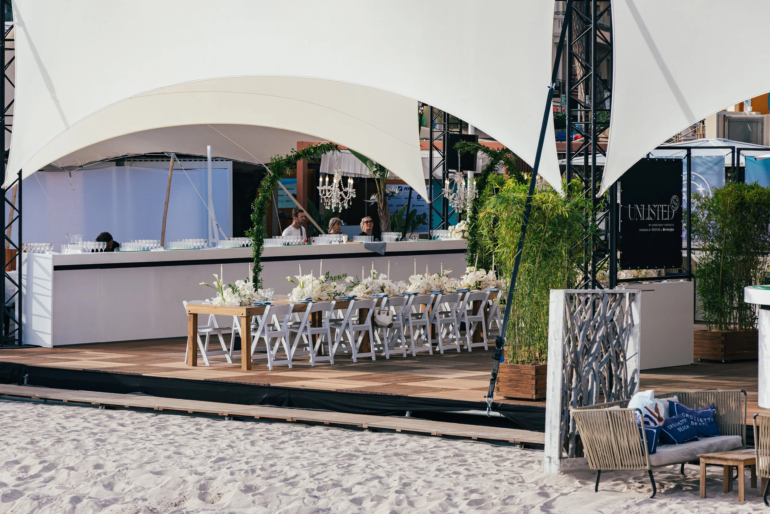 A beach wedding setup with a long table decorated with white flowers and candles, surrounded by white chairs under a white canopy, with a bar area and chandeliers in the background, and some lounge furniture with bags on the sand in the foreground.