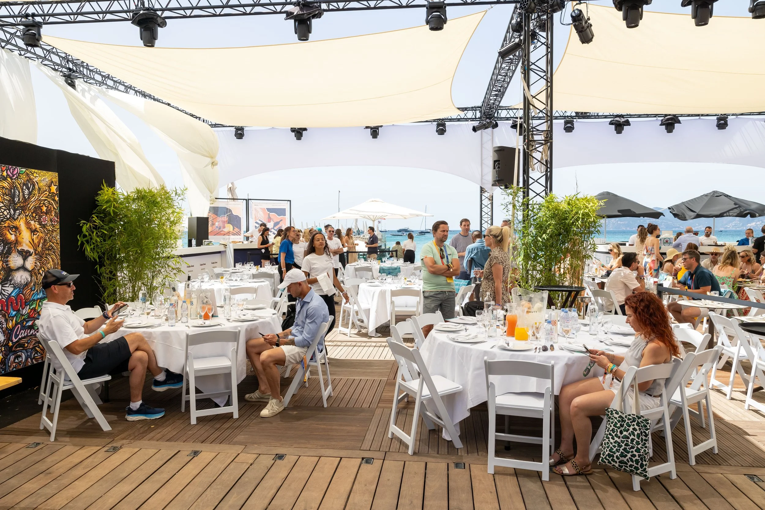 A lively outdoor dining area on a deck by the water with white tables and chairs, some guests sitting and others standing, under a large canopy. There are green plants, colorful artwork, and a view of boats and the ocean in the background.
