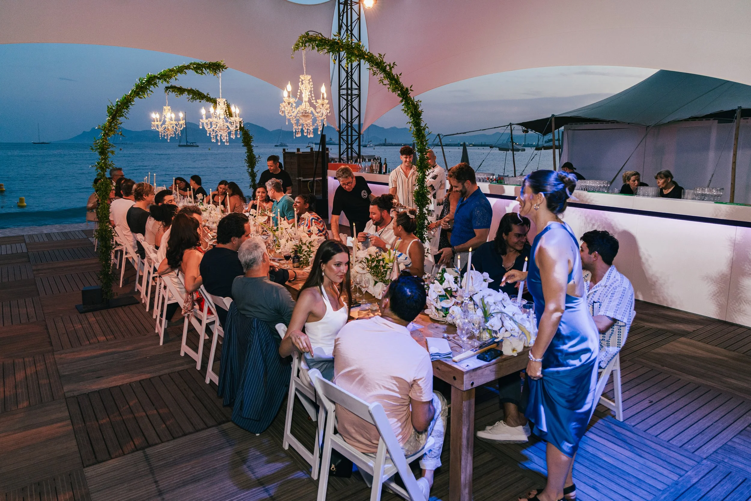 Outdoor dinner party at sunset with long table set with white flowers and candles, chandeliers hanging from a tent, and a waterfront view of boats and mountains in the background.