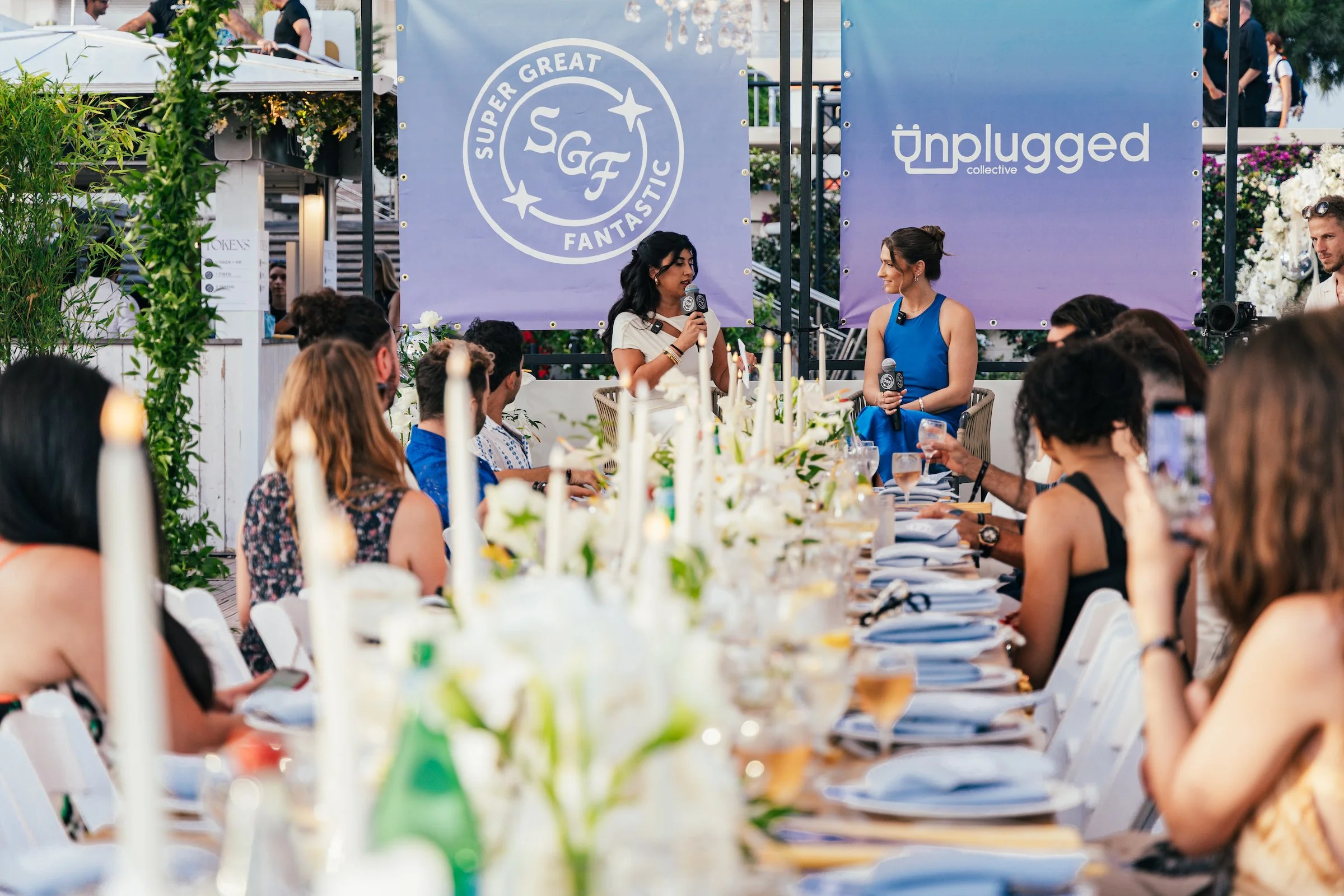 A woman in a white dress and a woman in a blue dress sitting on stage participating in a panel discussion during an outdoor event, with a audience seated at a long table decorated with candles and flowers, banners with logos behind them.