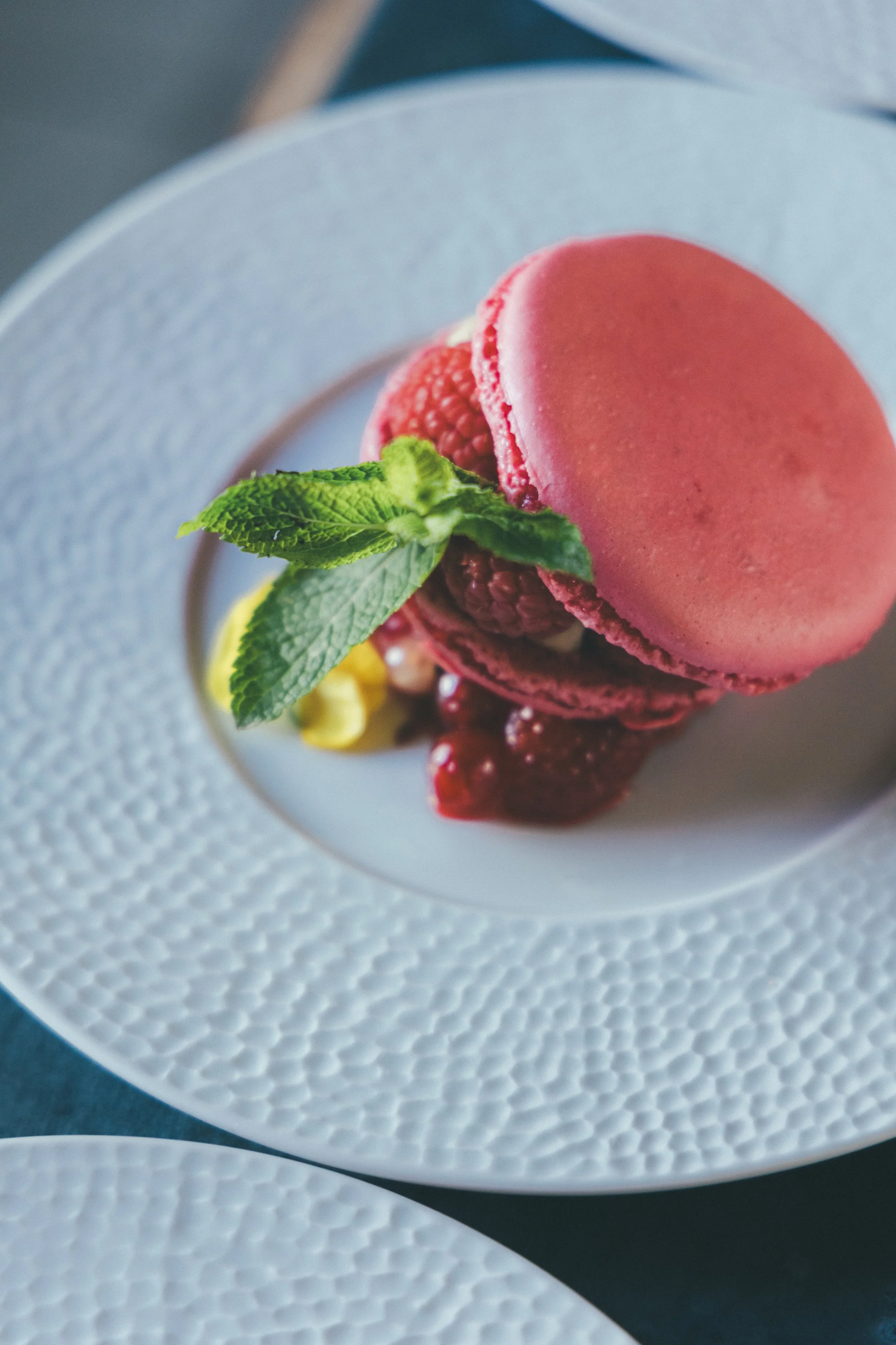 A plate with a pink macaron, fresh raspberries, and a sprig of mint.