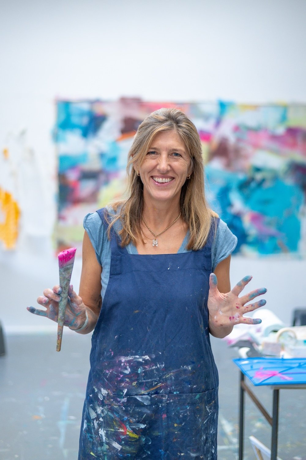 Abstract artist Trudie Tara Moulton with painted hands holding a paintbrush, smiling in an art studio with colourful abstract artwork in the background.
