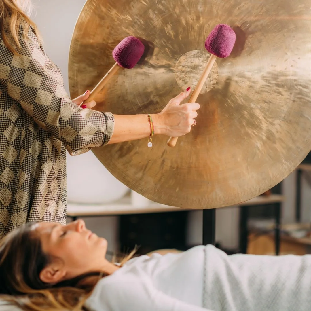 A woman reclines with eyes closed while a sound healer plays a wind gong with two mallets.