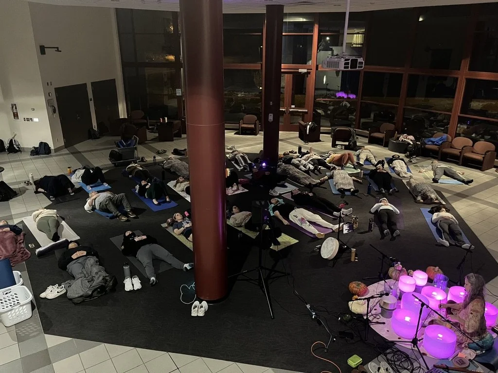 A large group of students are relaxing on yoga mats while a woman plays brightly lit crystal bowls at the front of the room.