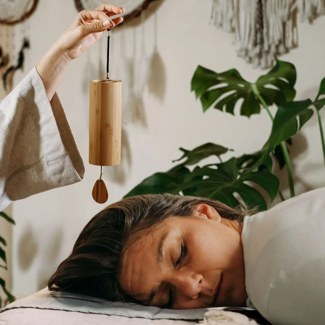 Photo of a woman lying down with eyes closed and a sound practitioner playing a koshi chime above her