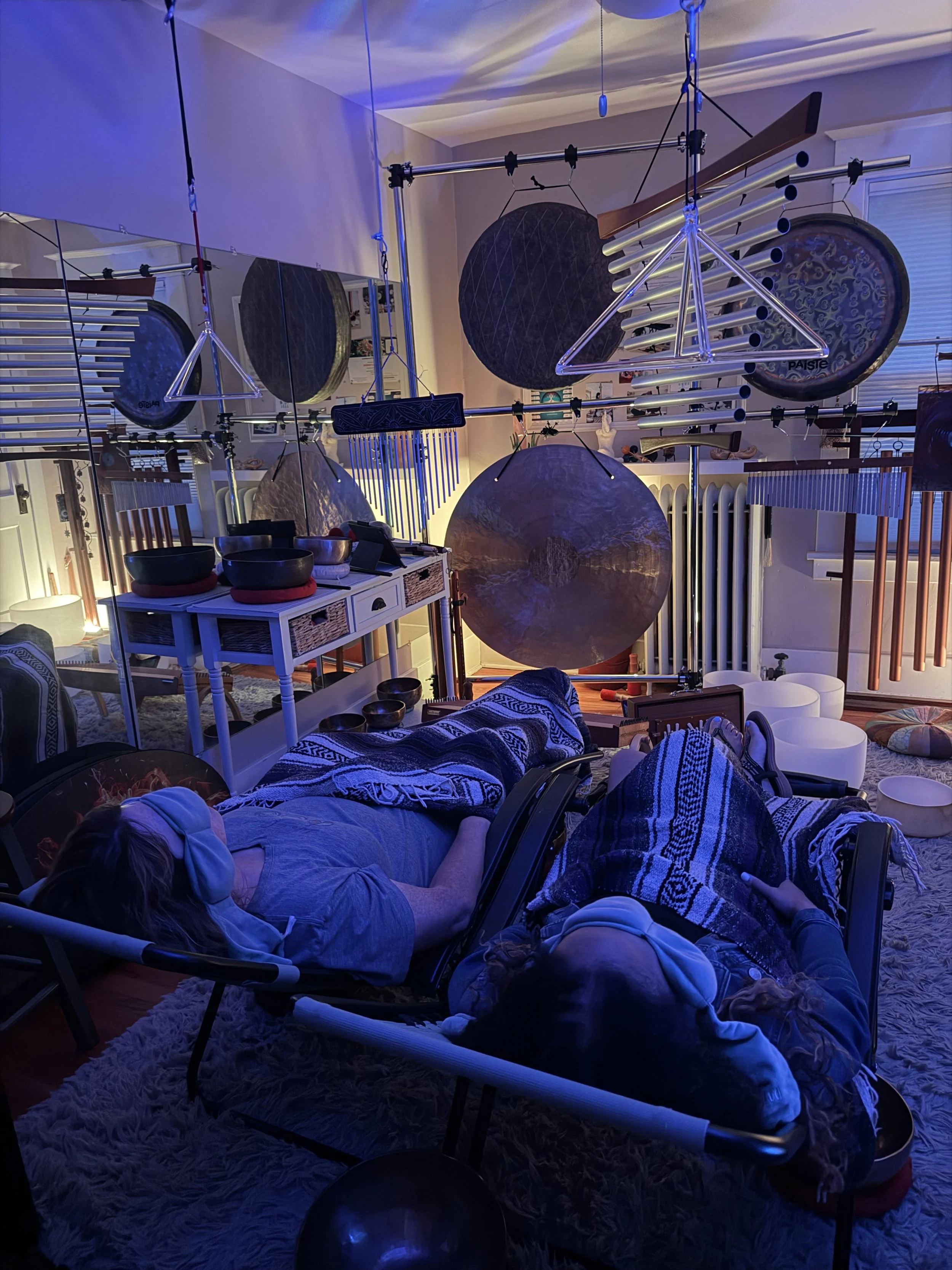 Two people re resting in zero-gravity chairs with gongs, bowls, chimes and more instruments all around them.
