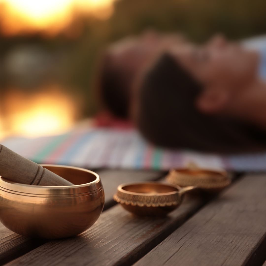 Outdoor image of small metal singing bowls in focus on a deck in the foreground with a couple relaxing on a blanket in the background