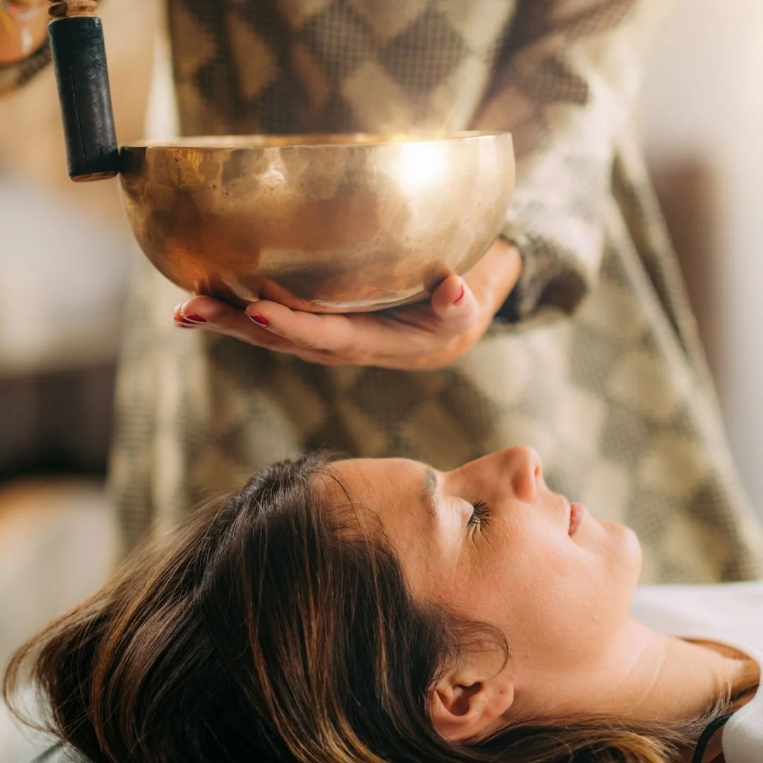 A woman reclines with eyes closed while a sound healer plays a Himalayan bowl above her head.