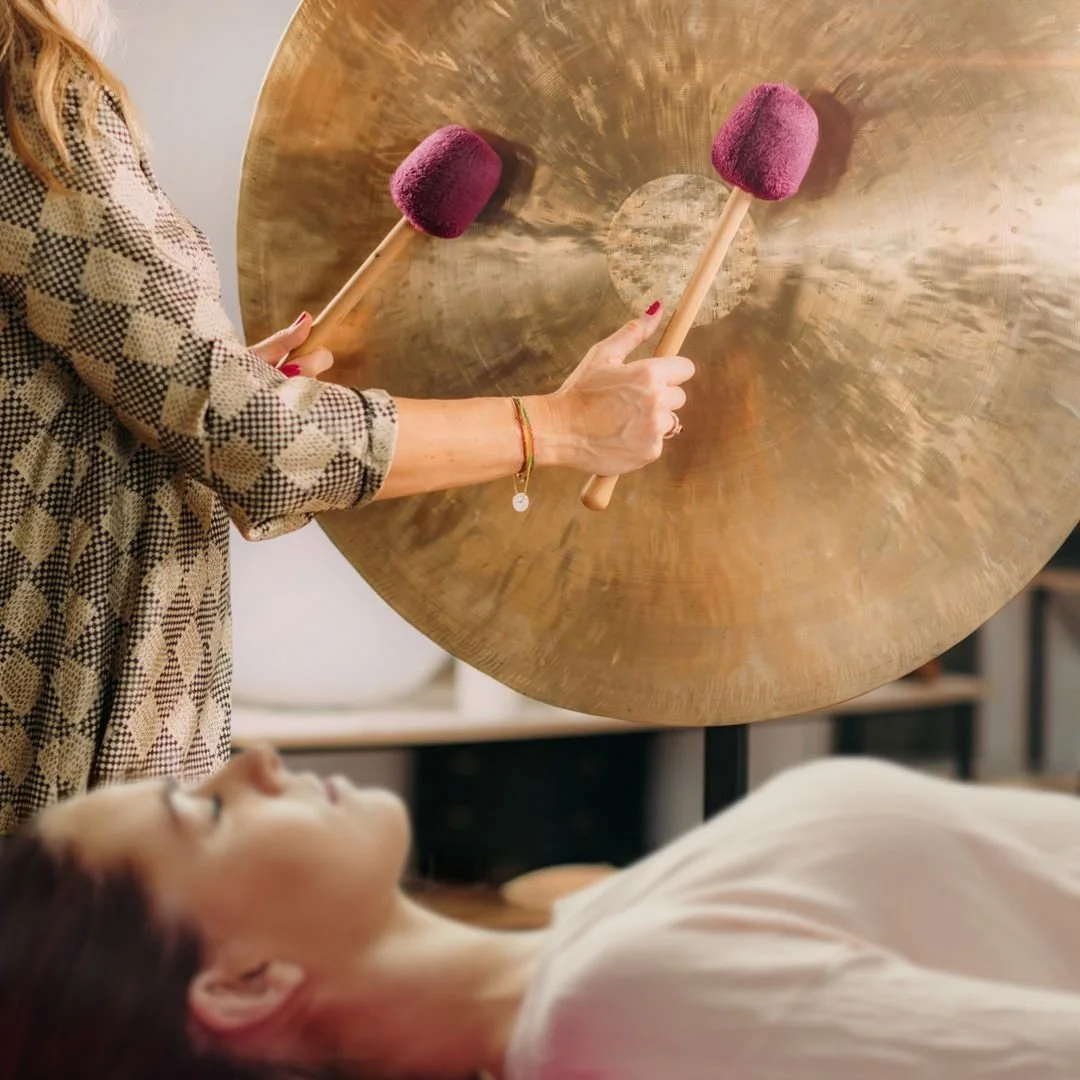 Photo of a woman lying down with eyes closed and a sound practitioner playing a gong