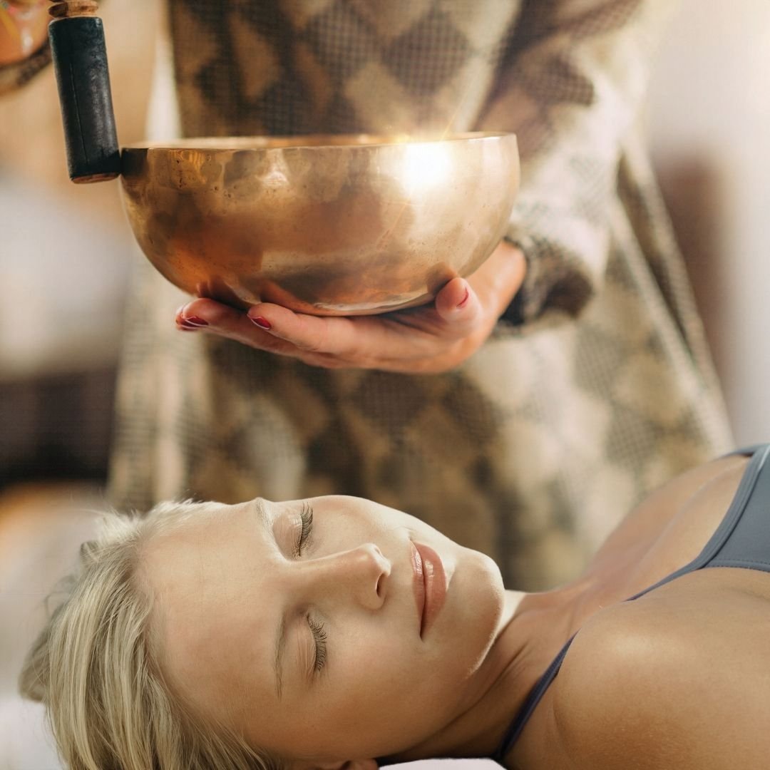 Photo of a woman lying down with eyes closed and a sound practitioner inviting a singing bowl above her