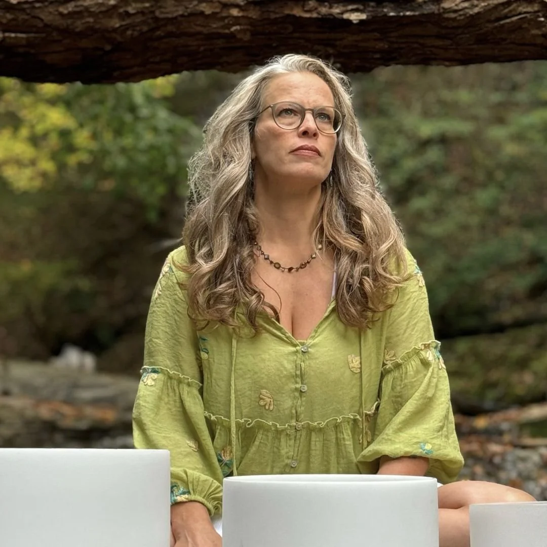 Nicholee Froese seated outdoors behind three crystal singing bowls, framed by trees, with a calm and steady expression.