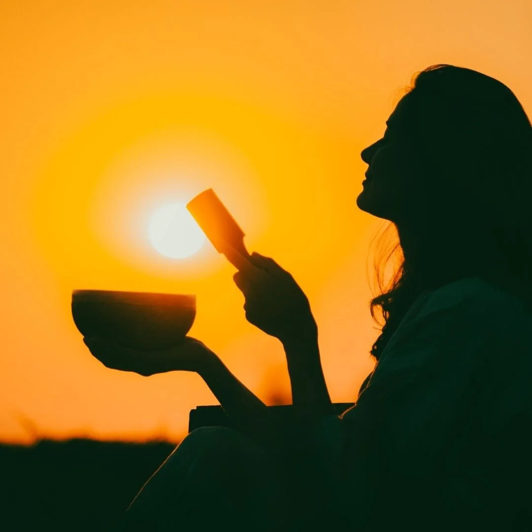 Silhouette of a woman holding a metal singing bowl and mallet in the setting sun