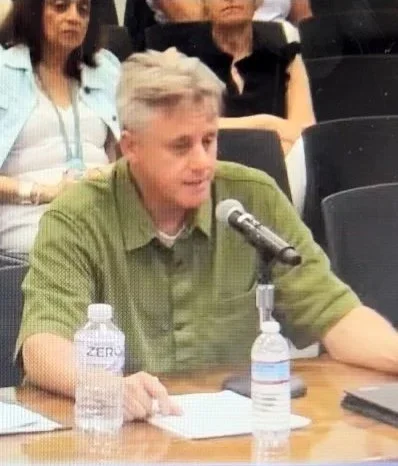 A man at a table equipped with water bottles and a microphone, indicating he is ready to speak or present.