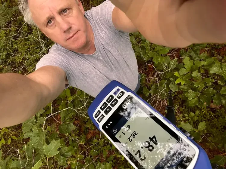 A man taking a selfie outdoors among green foliage, holding a GPS device displaying coordinates and altitude.