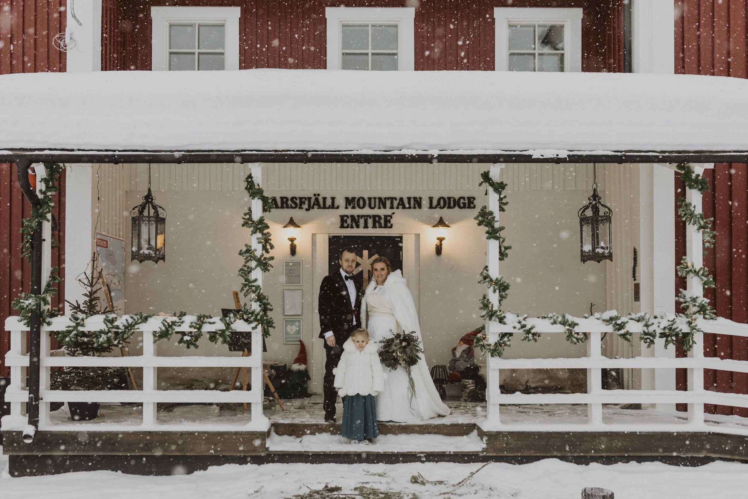 En bröllopsfotografering framför Marlsfjäll Mountain Lodge, snöfall, ett äkta par med ett barn, de är klädda i bröllops- och vinterkläder, dekorationer av granris och julklappar, utanför en rödmålad stuga.