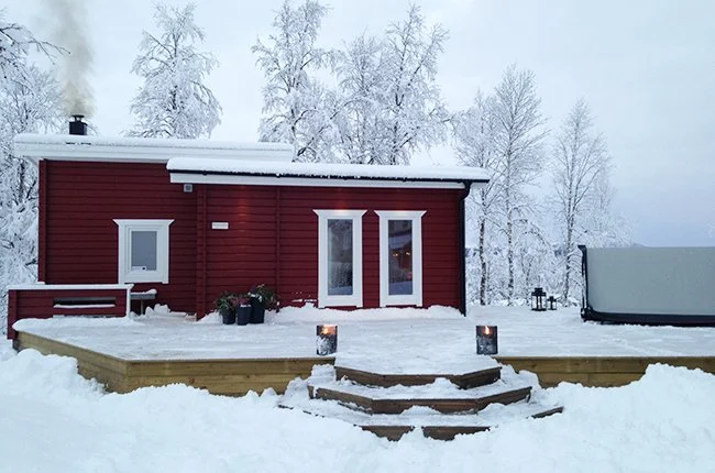 En röd liten stuga i snölandskap med trädörr och fönster, omgiven av snötäckta träd, trappor leder upp till entrén, uteplats med ljuslyktor och fågelholk.