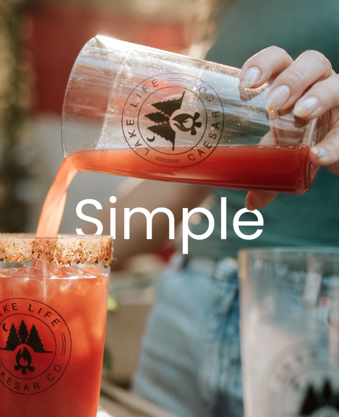 A person pouring a red beverage from a clear caesar glass with a logo, into another glass with a rim of chili powder, with the word 'simple' overlaid.