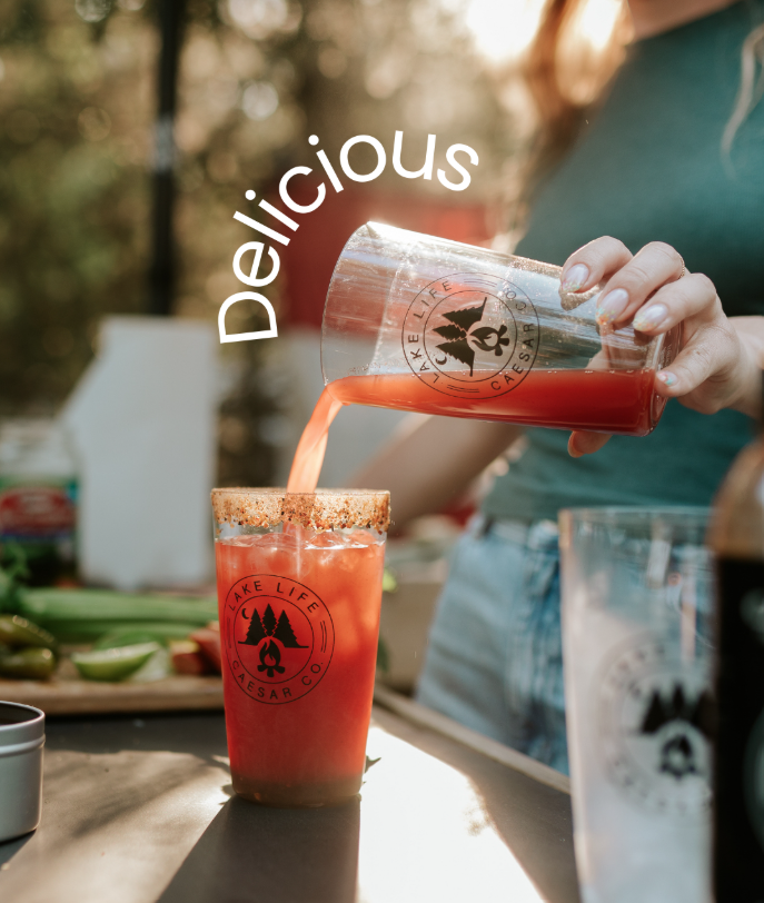 A person pouring a red drink into a glass with a rim of chili powder or seasoning outdoors, with the word 'Delicious' written beside the drink. Background includes lime wedges and other food items on a table.