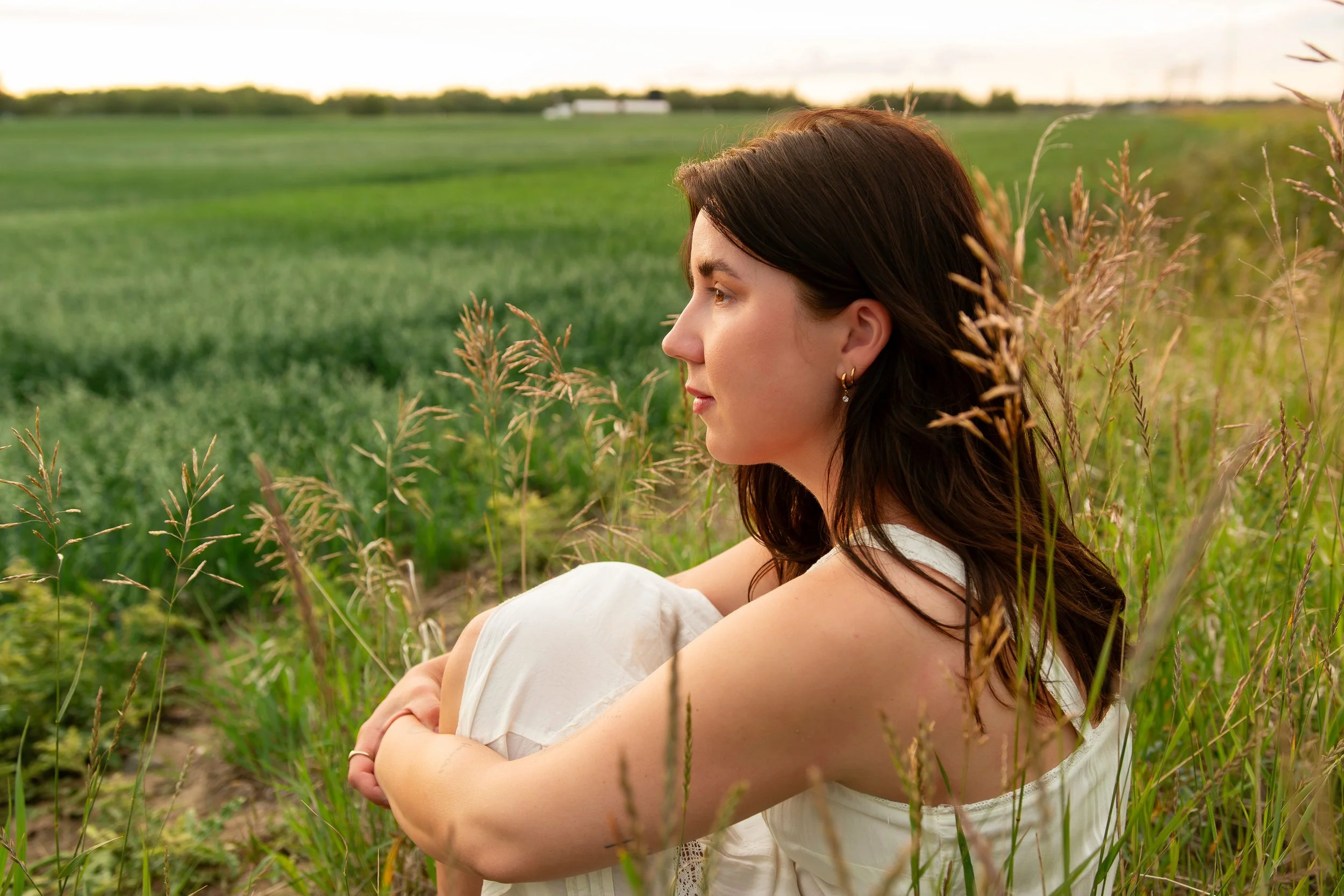 A woman with dark brown hair and earrings sitting in a field of tall grass, looking contemplatively into the distance during sunset.