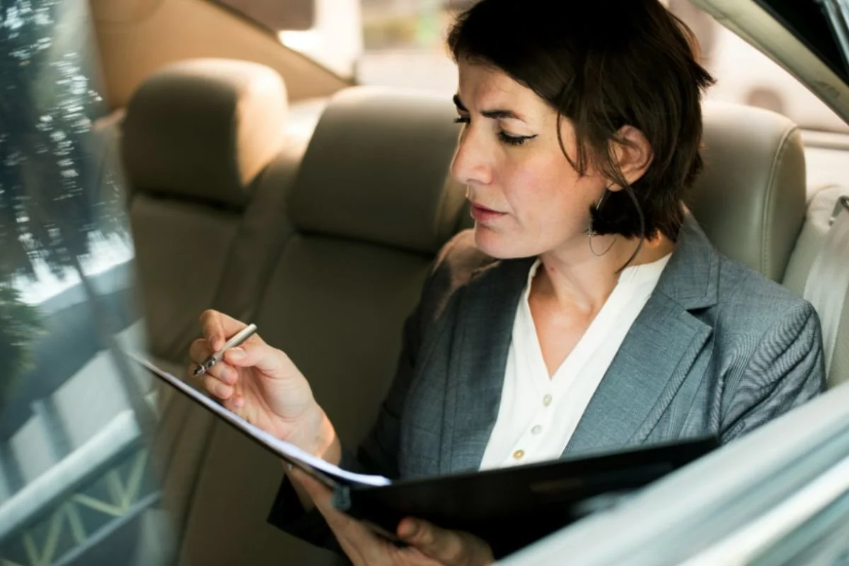 A professional woman in a blazer writing on a clipboard in the back of a car — depicting the nonstop decision-making demands that deplete cognitive capacity throughout the day
