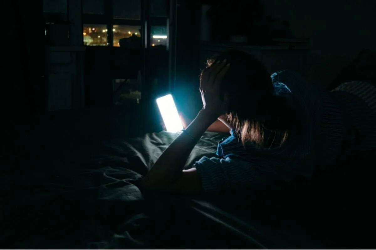 Woman lying in bed at night, face lit by her phone screen, one hand pressed to her head