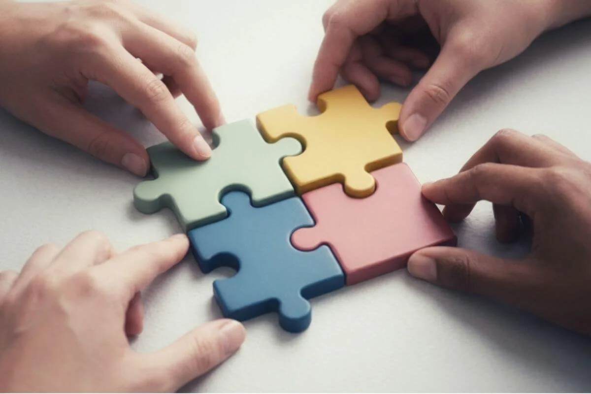 Four hands fit puzzle pieces together, each holding one part of a picture that only makes sense combined.