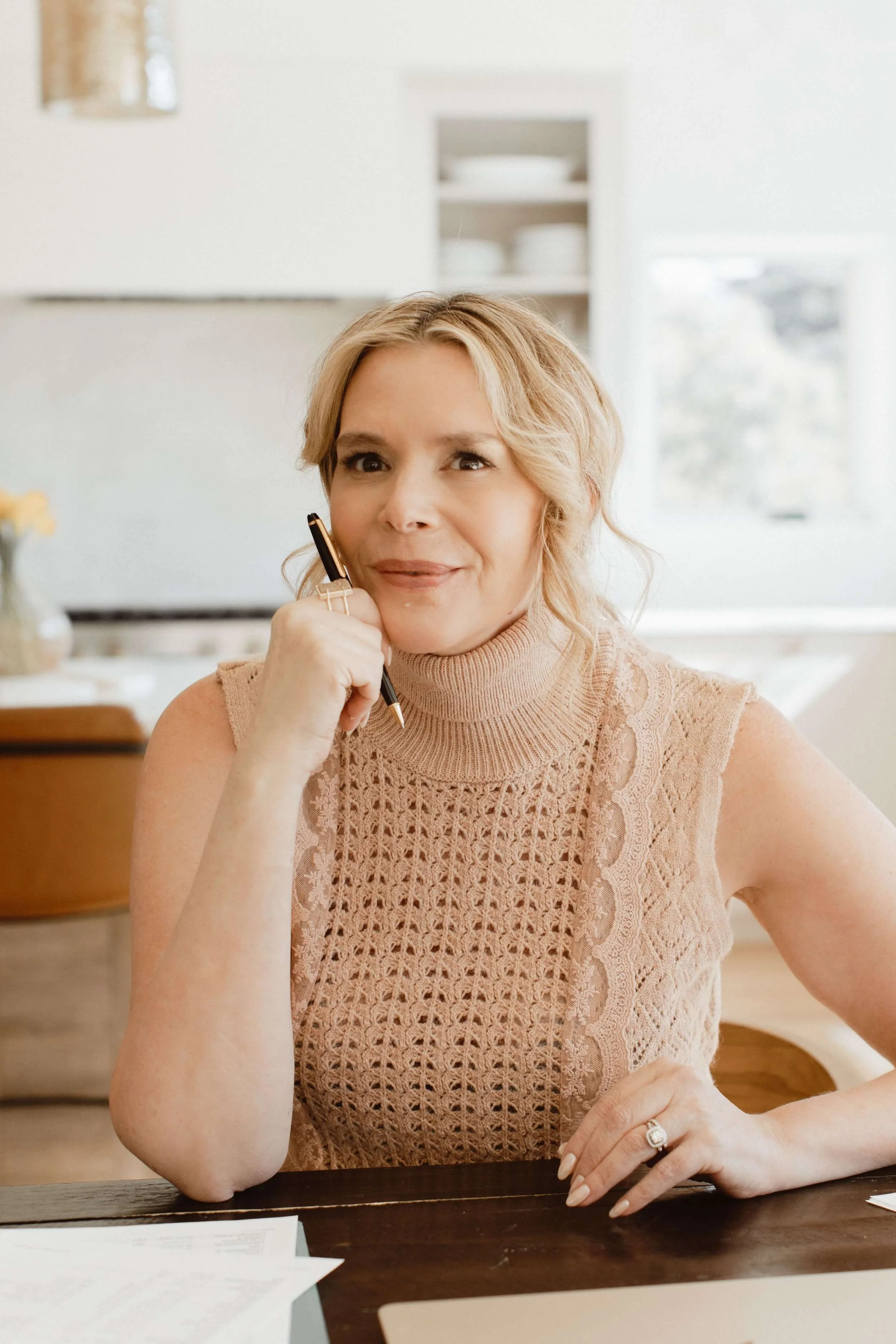 Dr. Kenna is wearing a peach-colored crochet sleeveless turtleneck top, holding a pen near her face, sitting at a table.