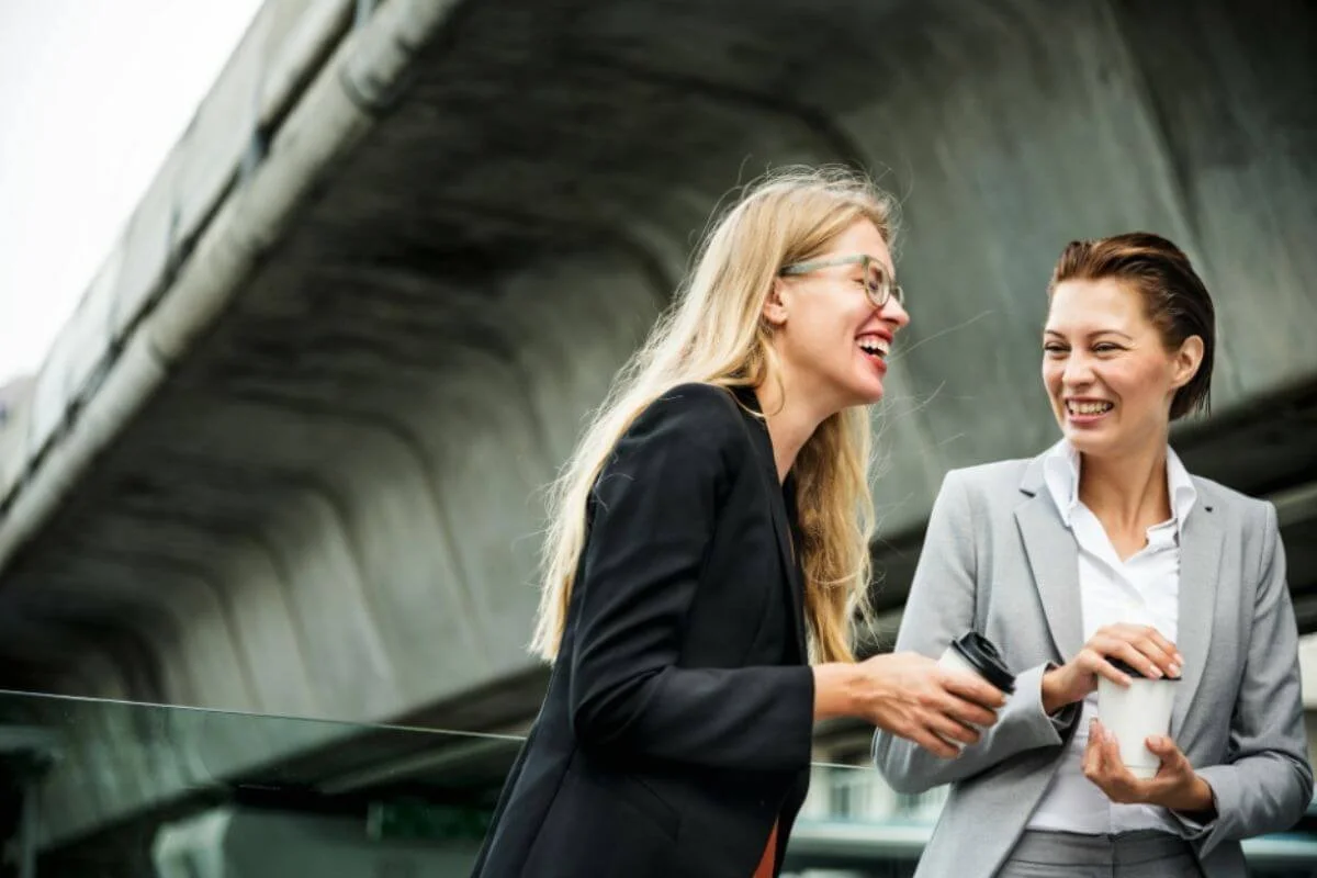 Two women in blazers laughing together over coffee before the workday starts