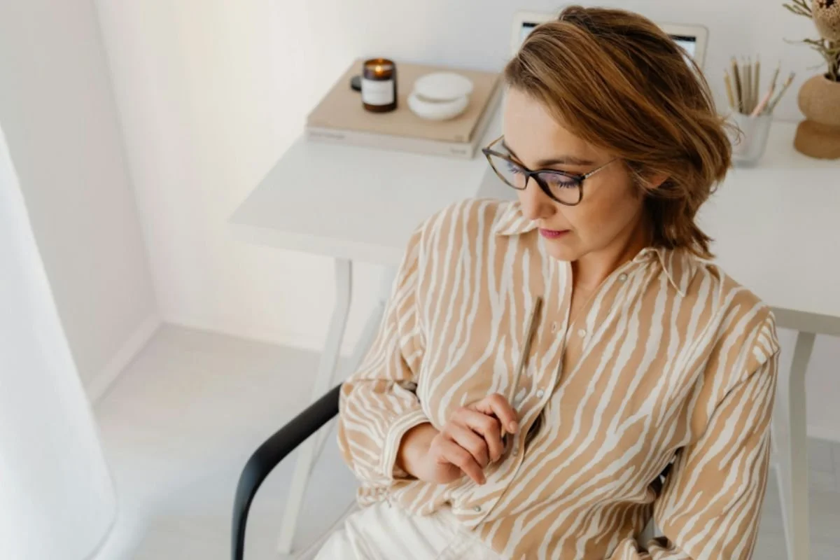 A high-performing woman with glasses sitting at her desk holding a pen, pausing in thought — representing the mental toll of chronic stress and nervous system overload