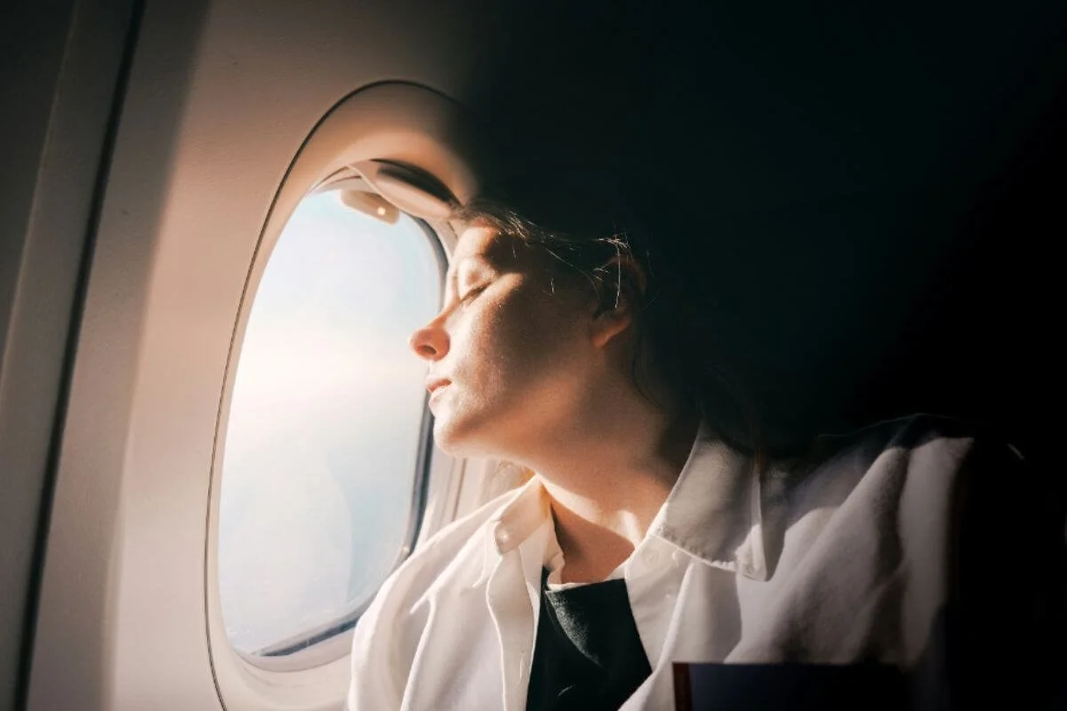 Woman with eyes closed against an airplane window, feeling the fatigue of jet lag