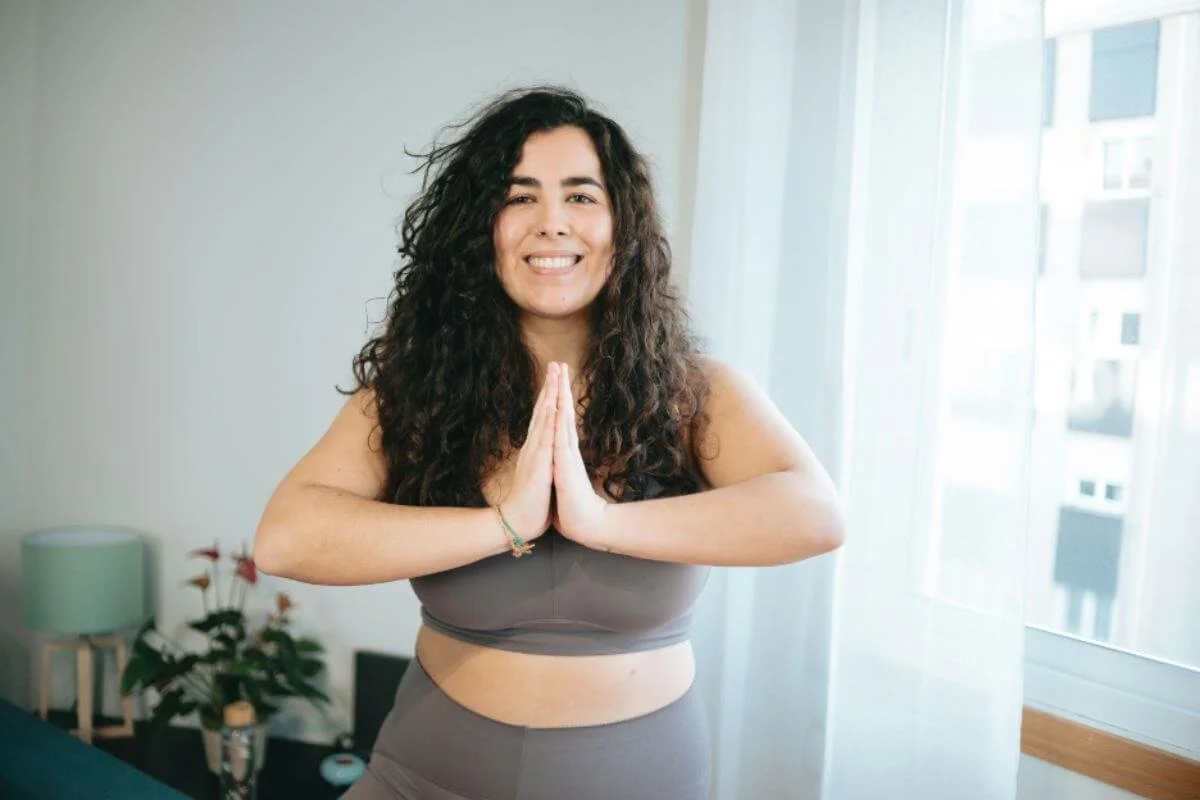 Woman in grey activewear standing in a sunlit room with hands pressed together in a yoga pose.