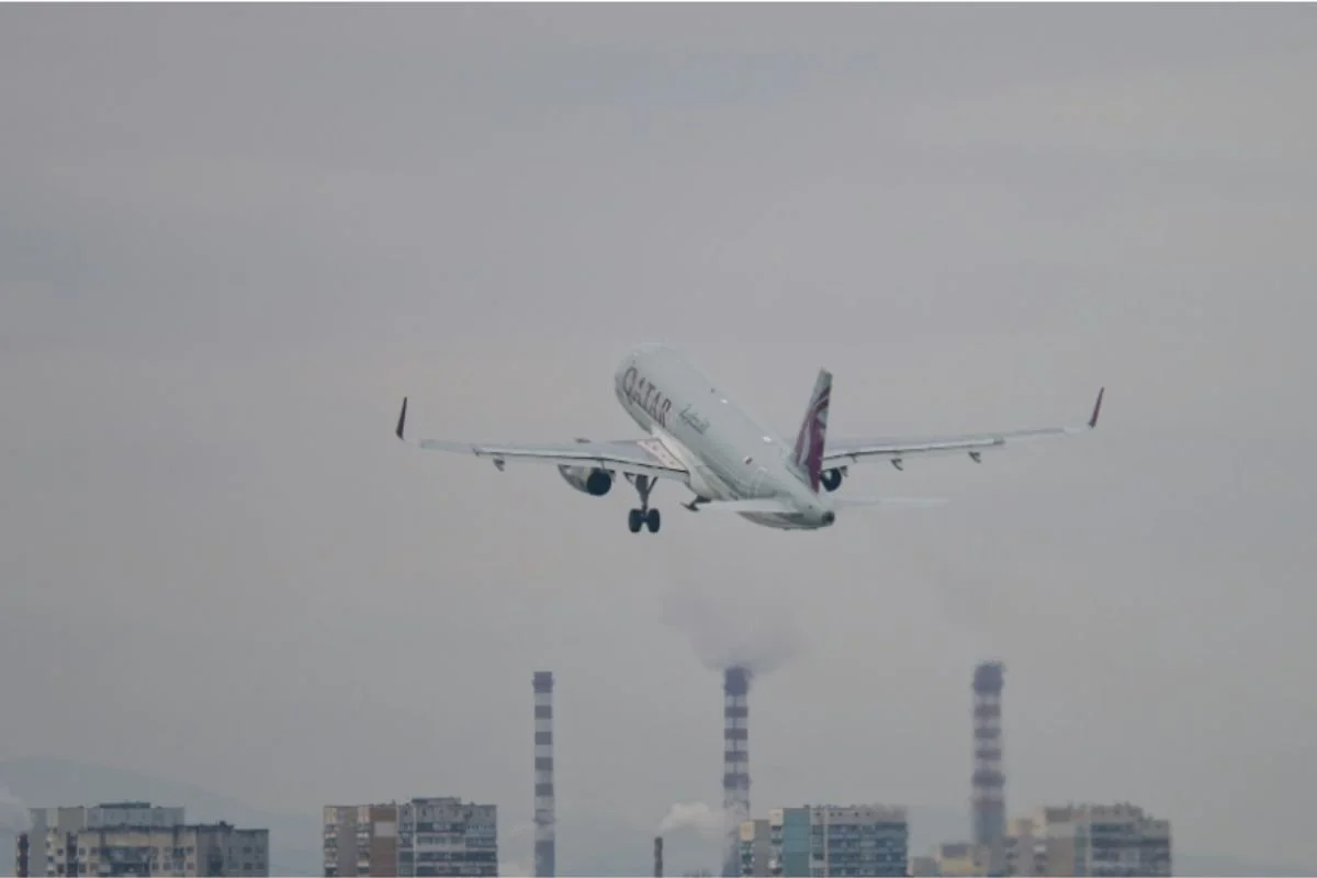 A plane climbing after takeoff, with smokestacks and hazy city buildings visible below.