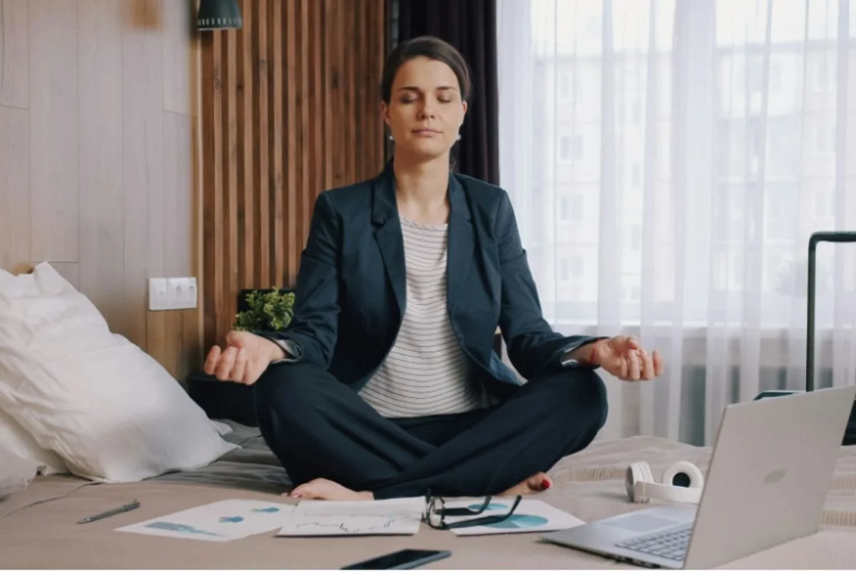 Businesswoman meditating cross-legged on a hotel bed, laptop and notes set aside beside her.