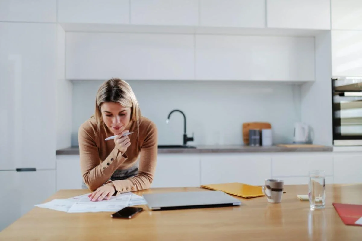 Woman reviewing paperwork at a kitchen counter with a laptop and coffee nearby