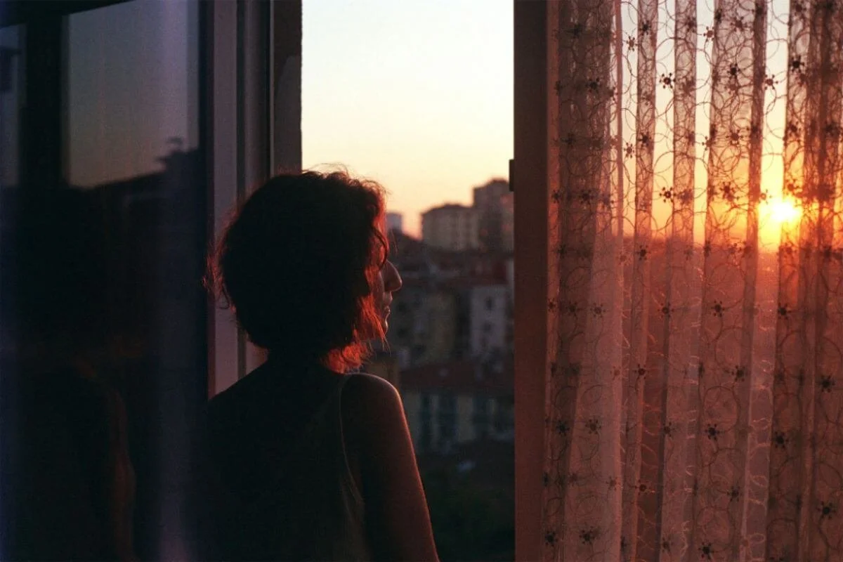 Woman gazing at a city sunset through a hotel window, her circadian rhythm out of sync with local time