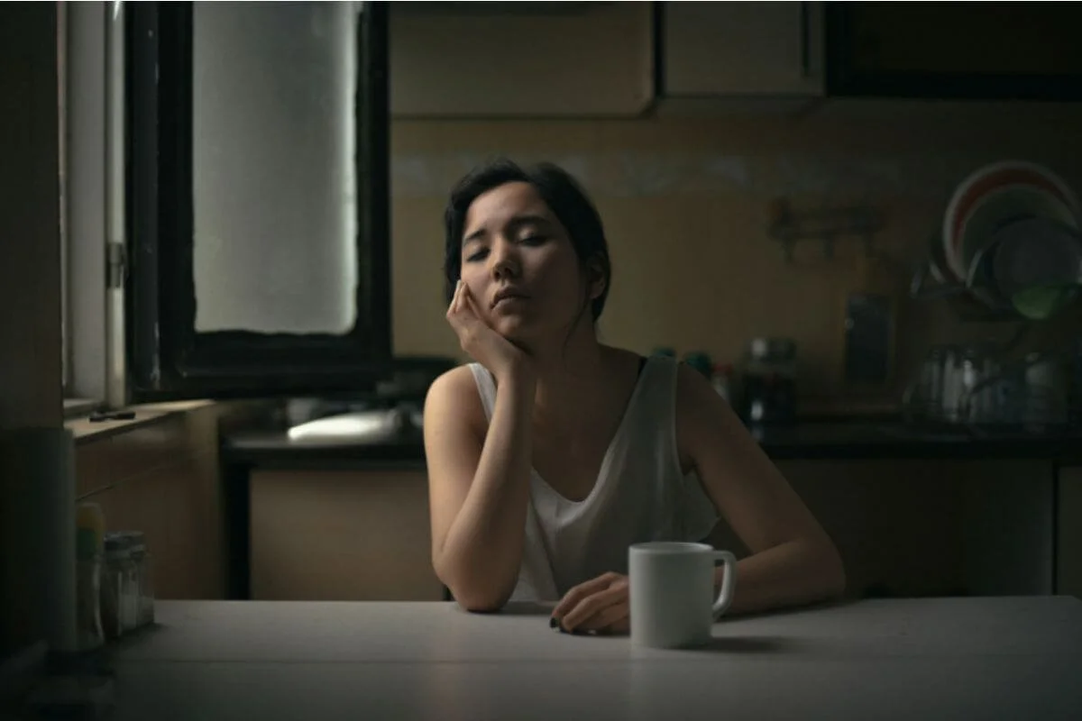 Tired woman resting her head in her hand at a kitchen table beside a coffee mug