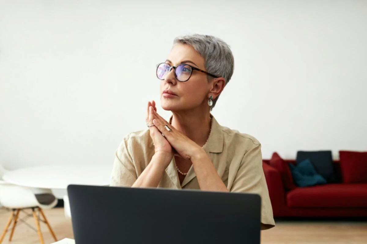 Woman with hands clasped in thought at her laptop, considering the connection between chronic stress and hormonal changes