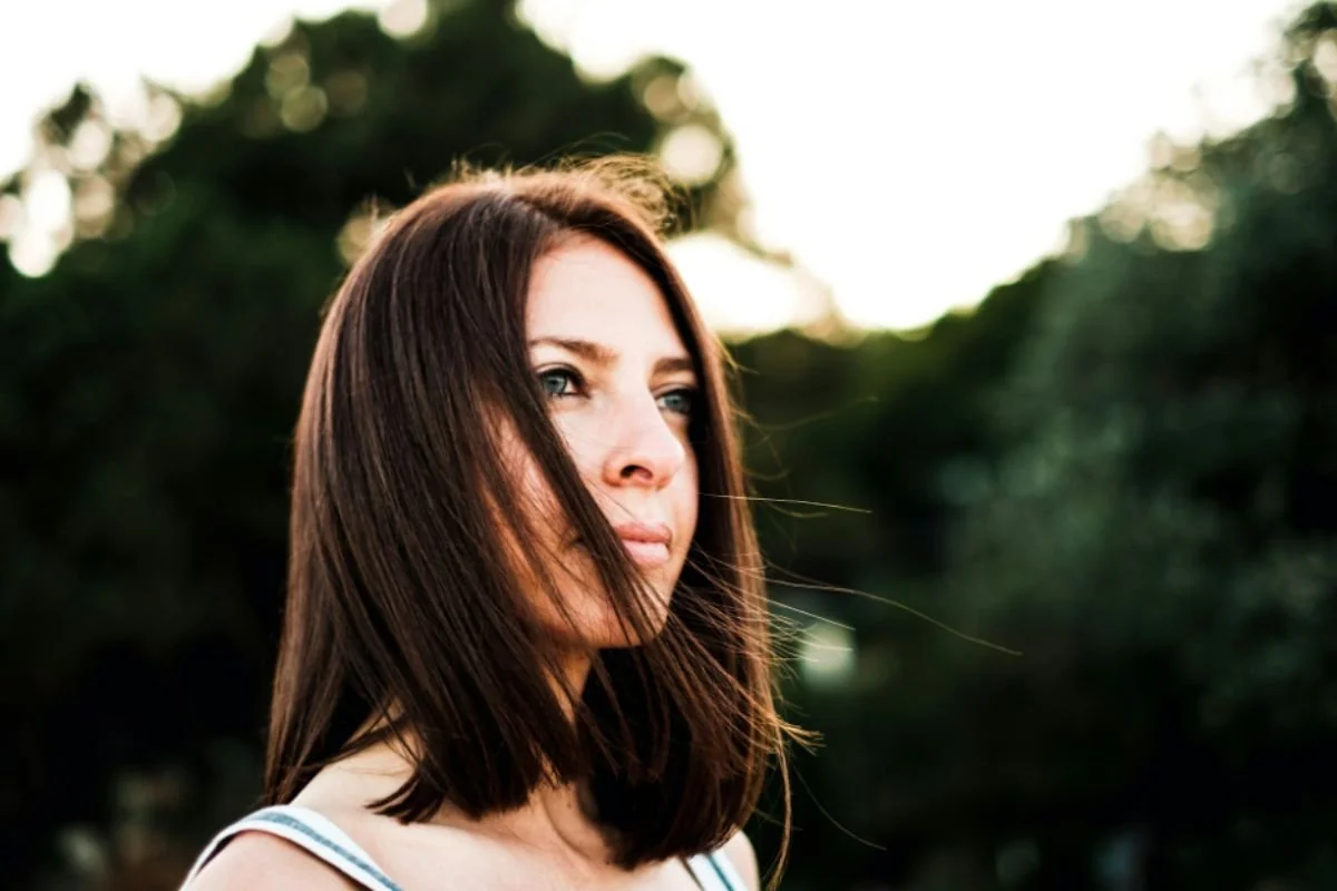 A woman standing outdoors with wind blowing her hair across her face, looking into the distance — capturing the quiet exhaustion and reflection that comes with carrying too much for too long