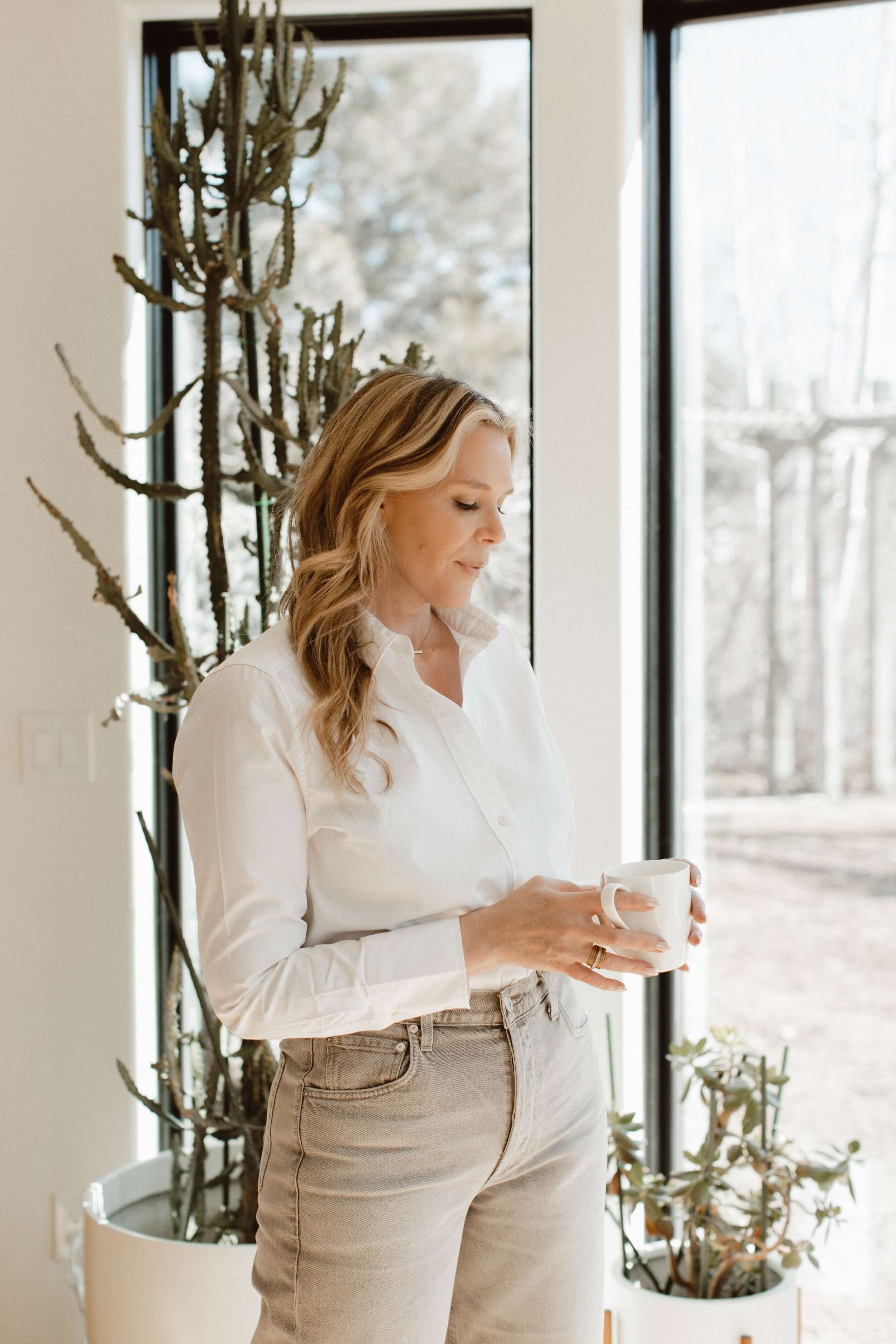 Dr. Kenna is wearing a white shirt and beige pants, holding a white mug, standing near a large window with a cactus plant and other greenery in a white pot behind her.