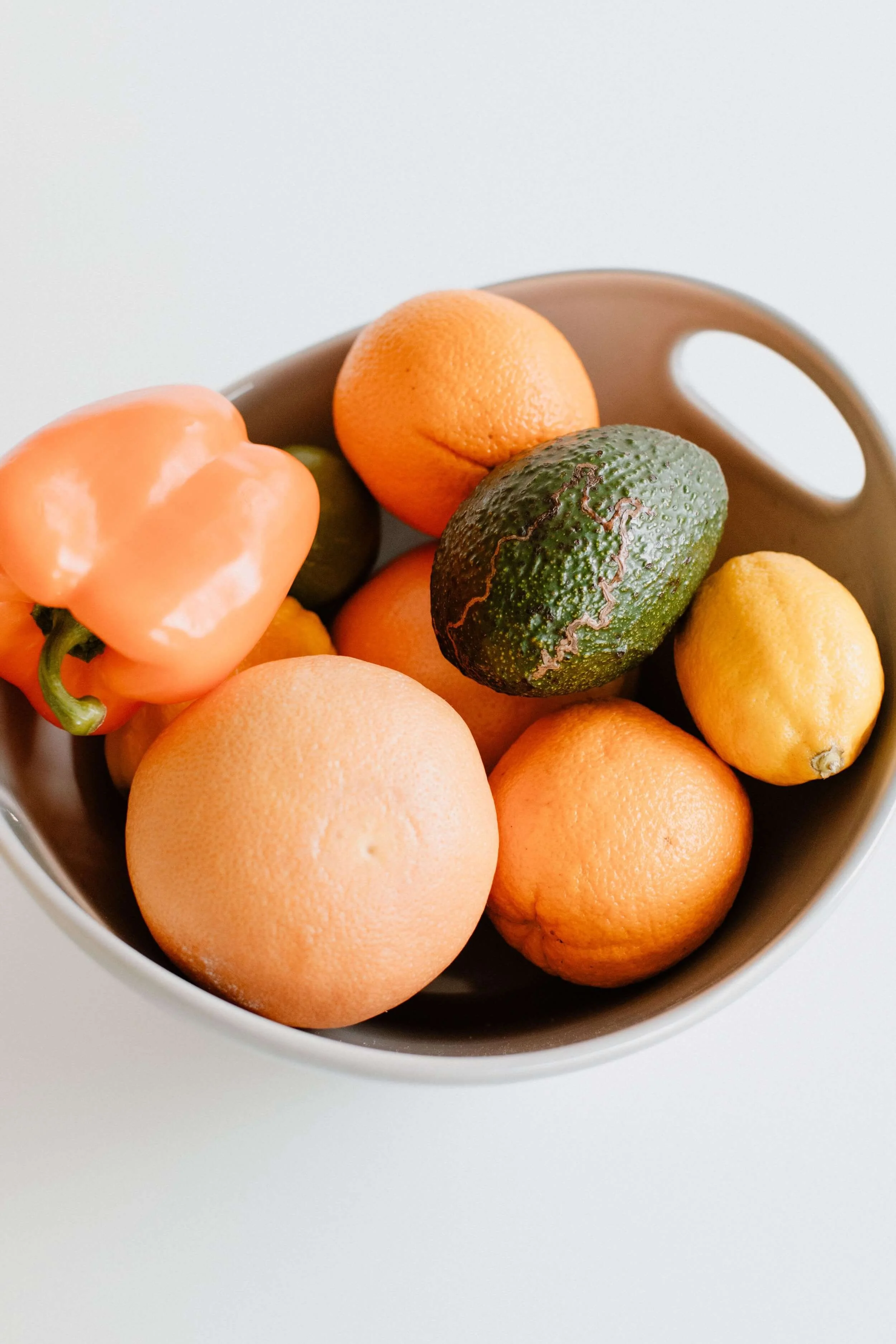 A bowl filled with various fruits including an orange, a lemon, an avocado, a grapefruit, a yellow bell pepper, and a small green lime.