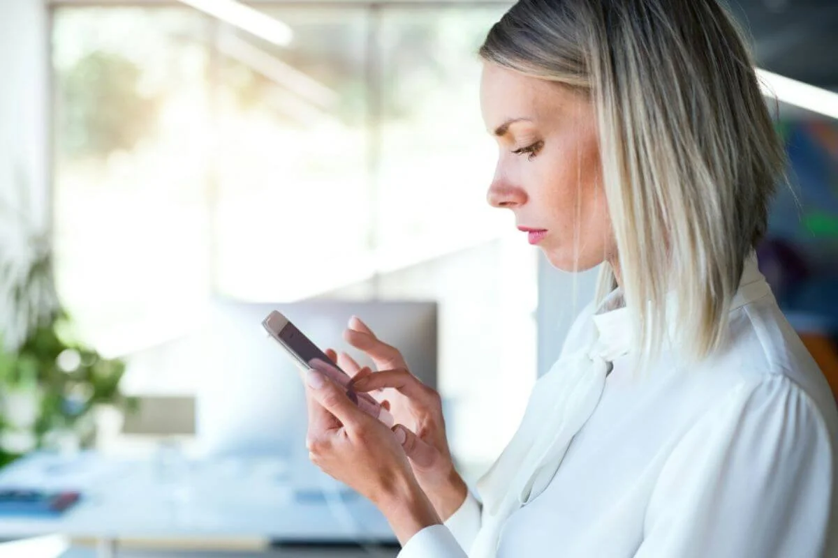 Woman in a white blouse looking down at her phone in a bright office with plants in the background.
