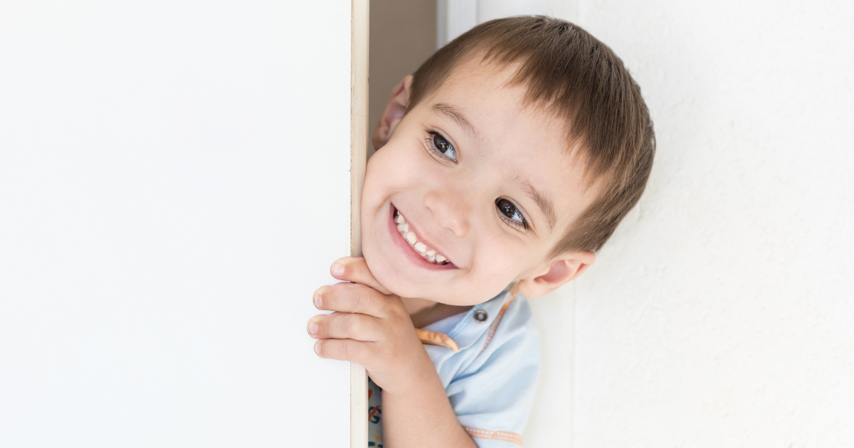 Smiling toddler boy peeking out from behind a bedroom door during toddler sleep training for separation anxiety
