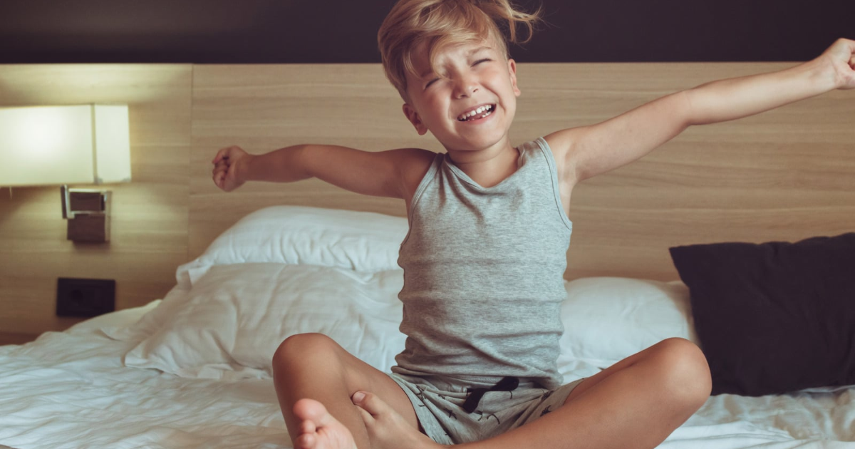 Tired child stretching and yawning while sitting on a bed early in the morning.