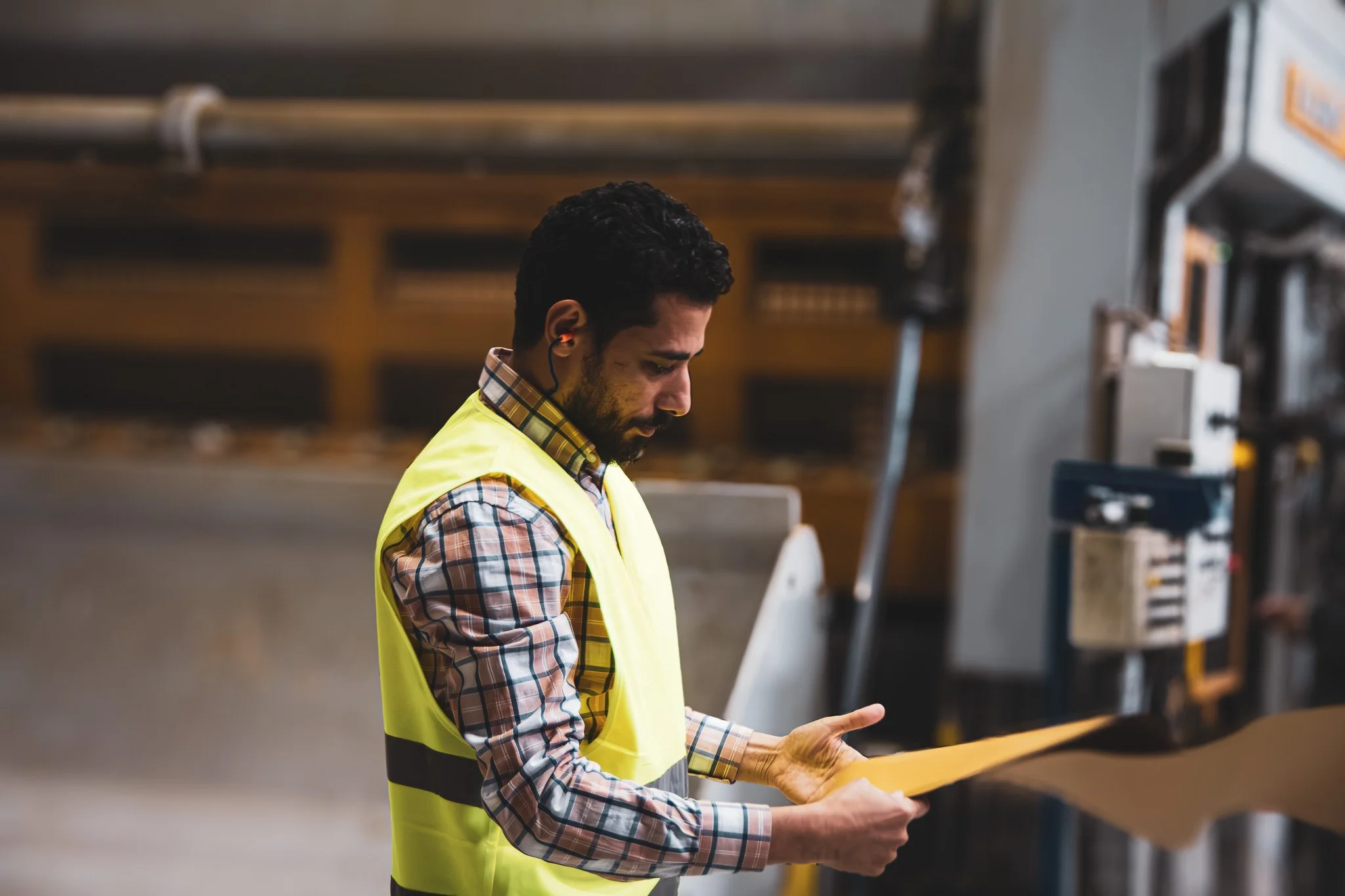 Man wearing a yellow safety vest and plaid shirt inspecting a document in a warehouse.