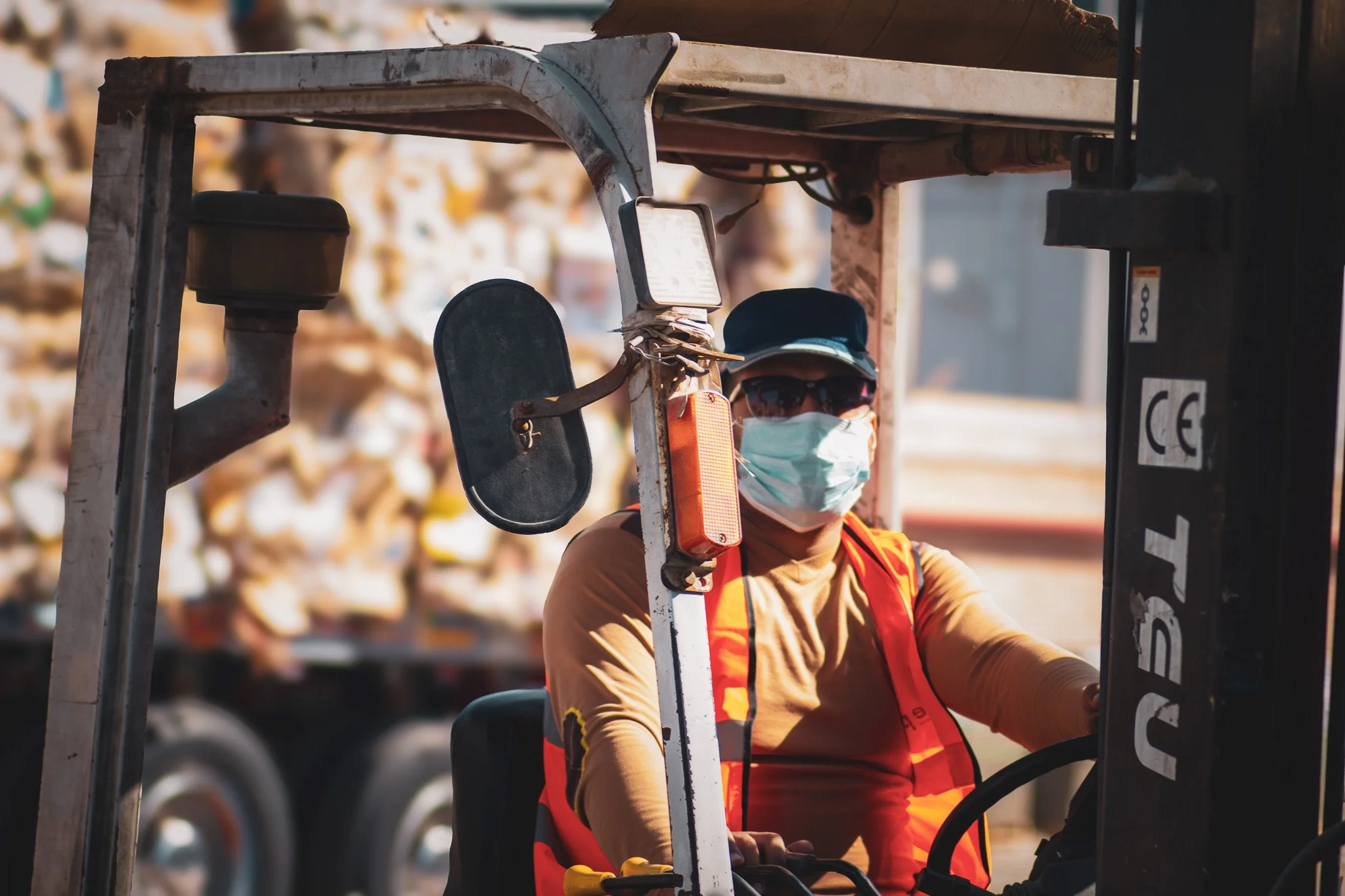 A man wearing a face mask, sunglasses, and a cap driving a forklift truck. The man has a reflective orange safety vest and is working outdoors.
