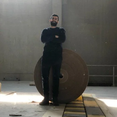 A man with a beard and glasses wearing a black sweater and pants, standing with arms crossed in front of a large industrial wheel inside a warehouse.