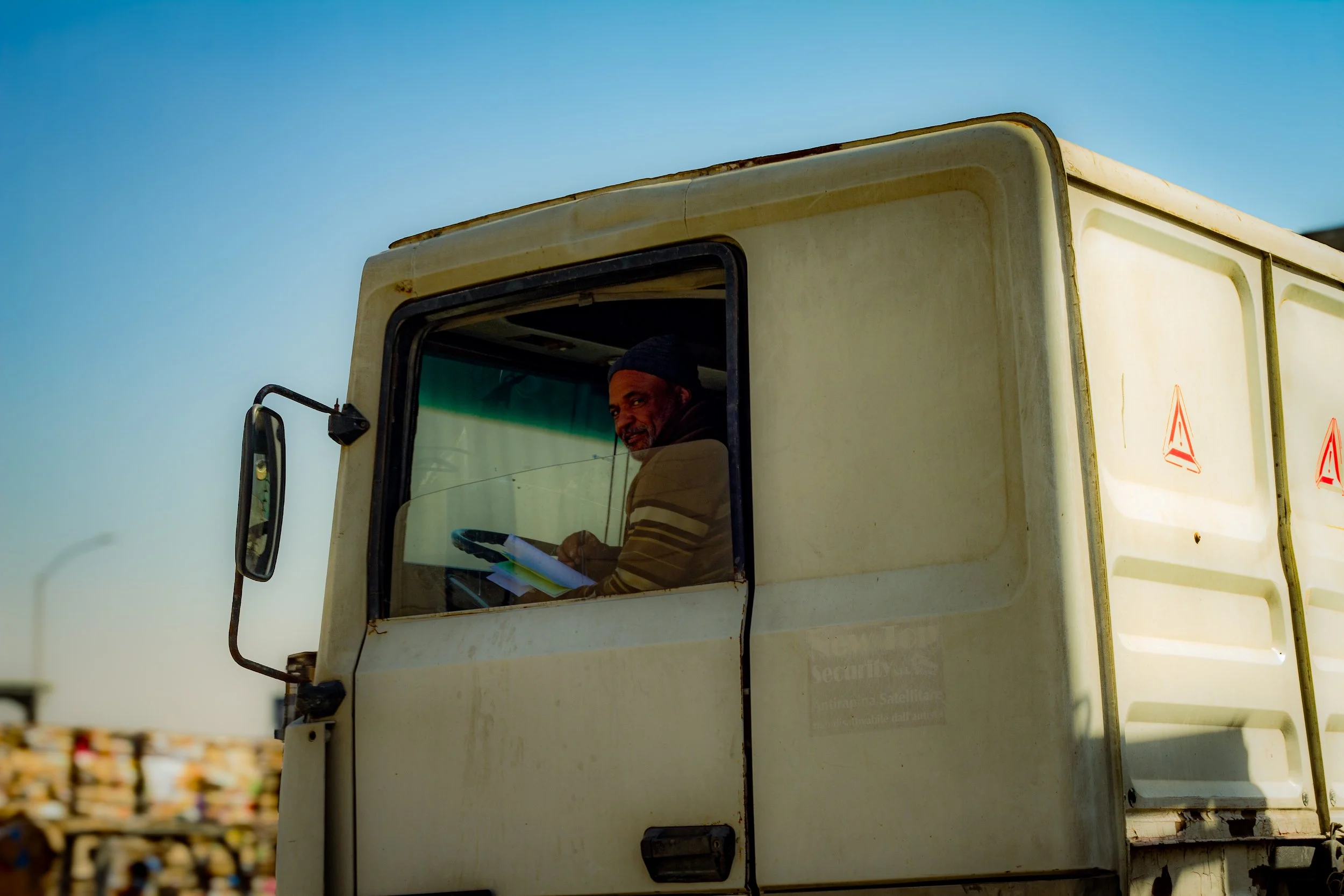 A man in a beanie sitting inside the cabin of a large truck, looking out the window and holding a paper or a notebook, with a clear blue sky in the background.